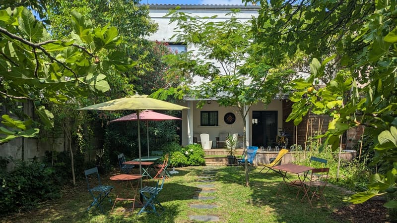 Jardin arboré avec tables colorées, parasols et pas japonais devant la terrasse d'un bâtiment